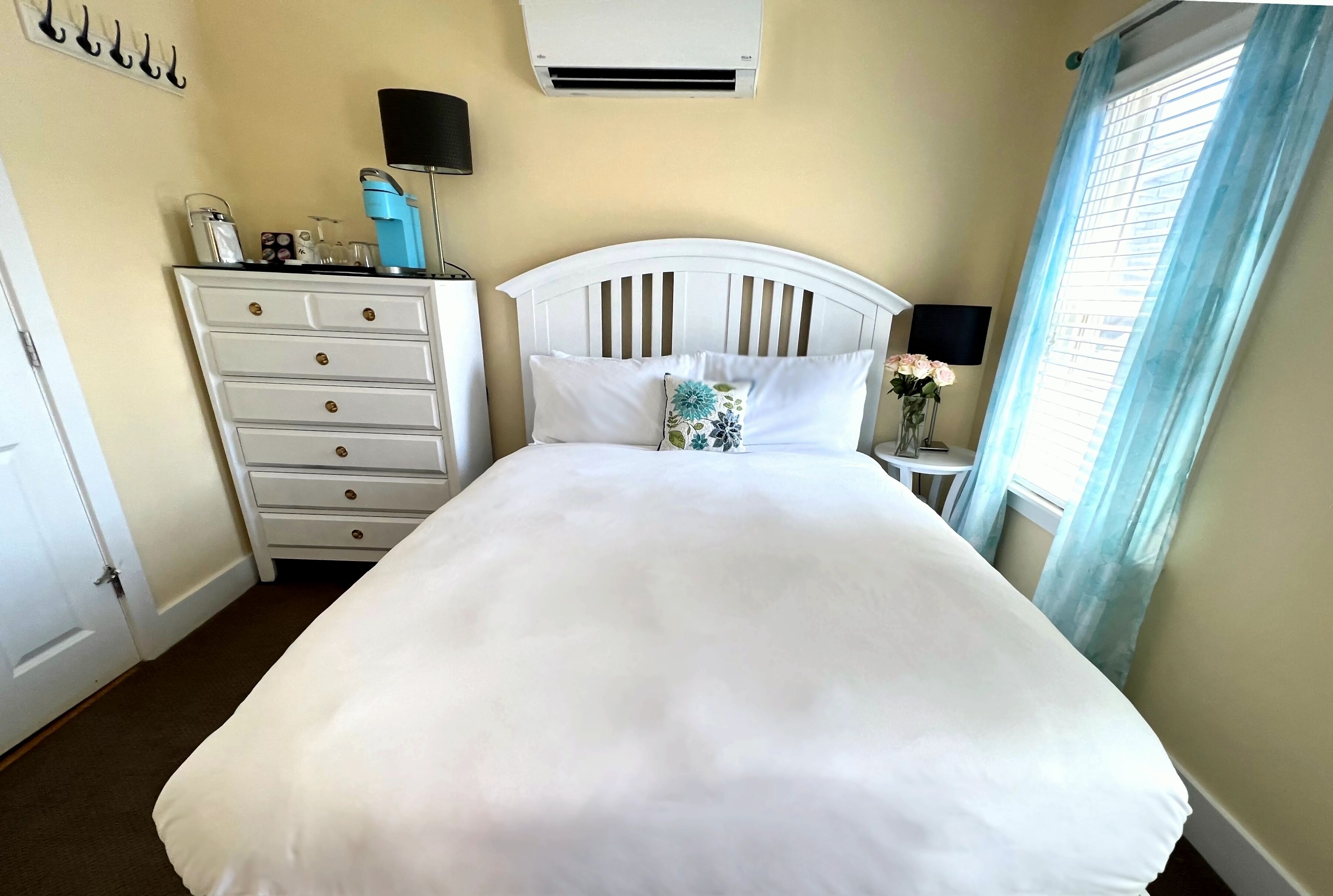 A neatly made bed with white bedding, a white dresser with drawers, a bedside table with a lamp, and a window with light blue curtains.
