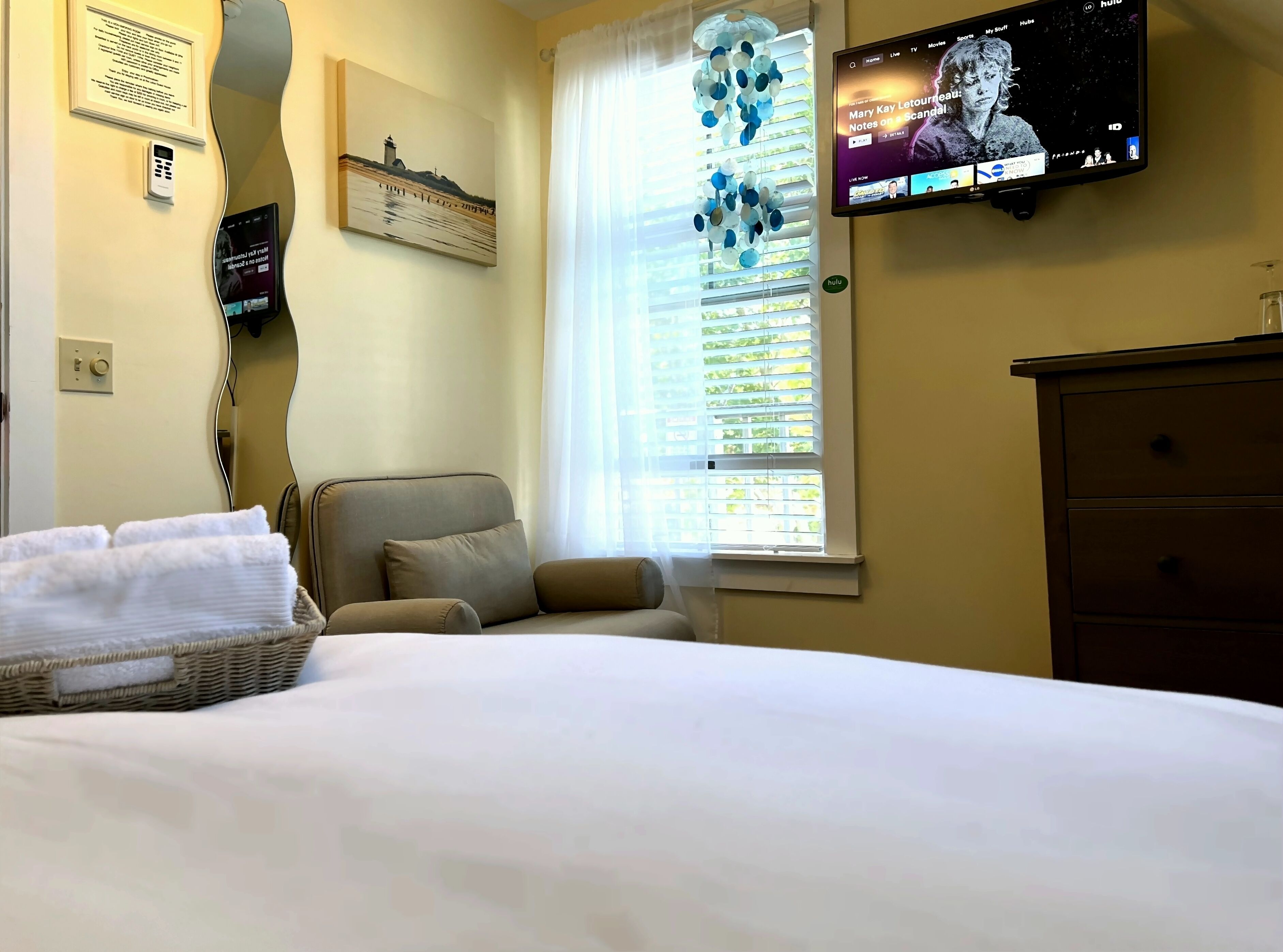 Une chambre d’hôtel avec un lit, une chaise, un téléviseur fixé au mur, une fenêtre avec des stores et un thermostat mural.