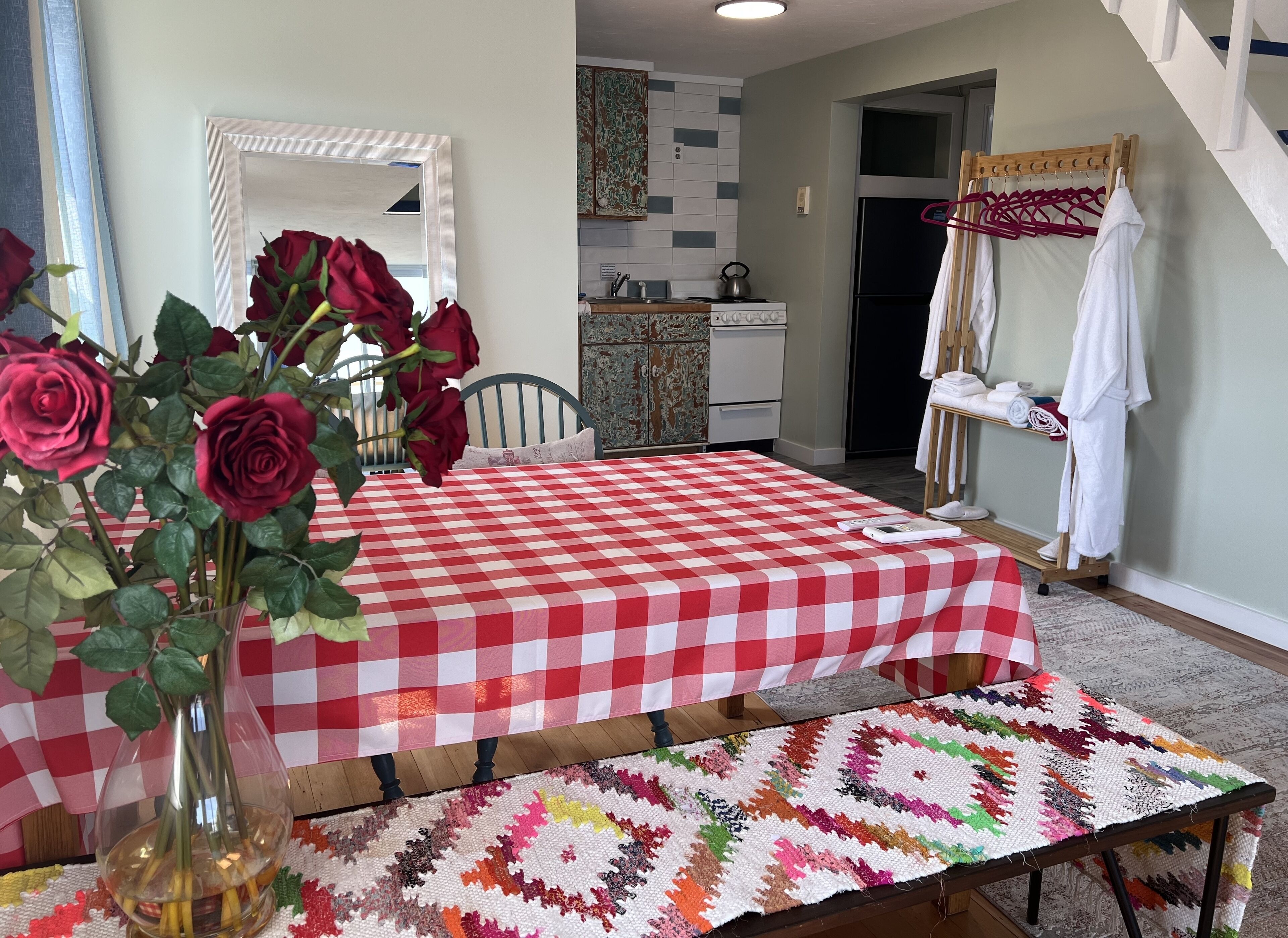 Un comedor con mantel a cuadros, un jarrón de rosas, un camino de mesa tejido y colorido, y una cocina al fondo.