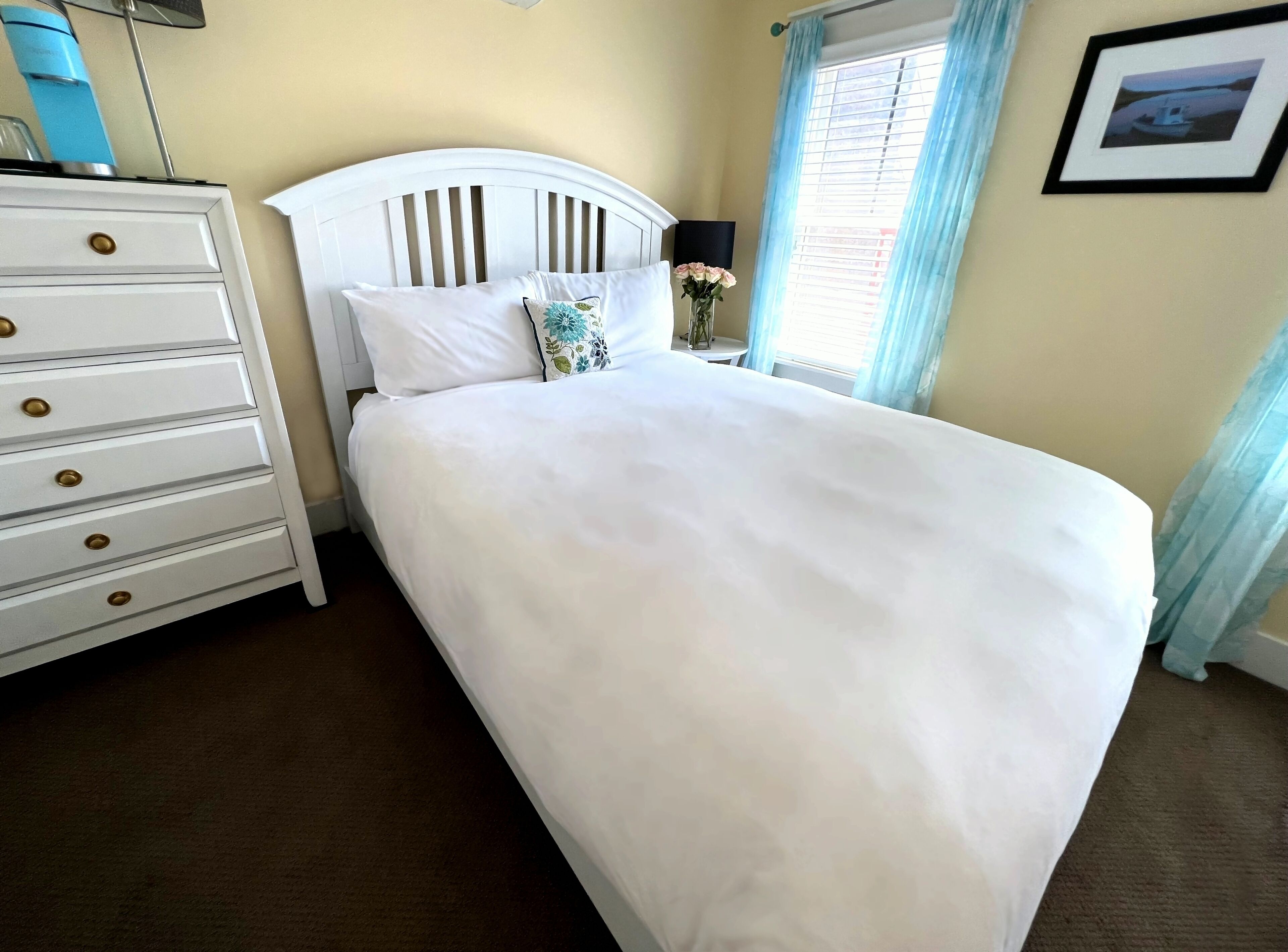 Uma cama bem arrumada com roupa de cama branca, um penteado branco com puxadores dourados e uma pequena mesa com abajur e flores.