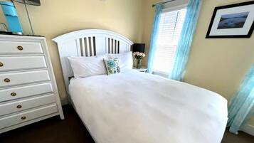 Uma cama bem arrumada com roupa de cama branca, um penteado branco com puxadores dourados e uma pequena mesa com abajur e flores.