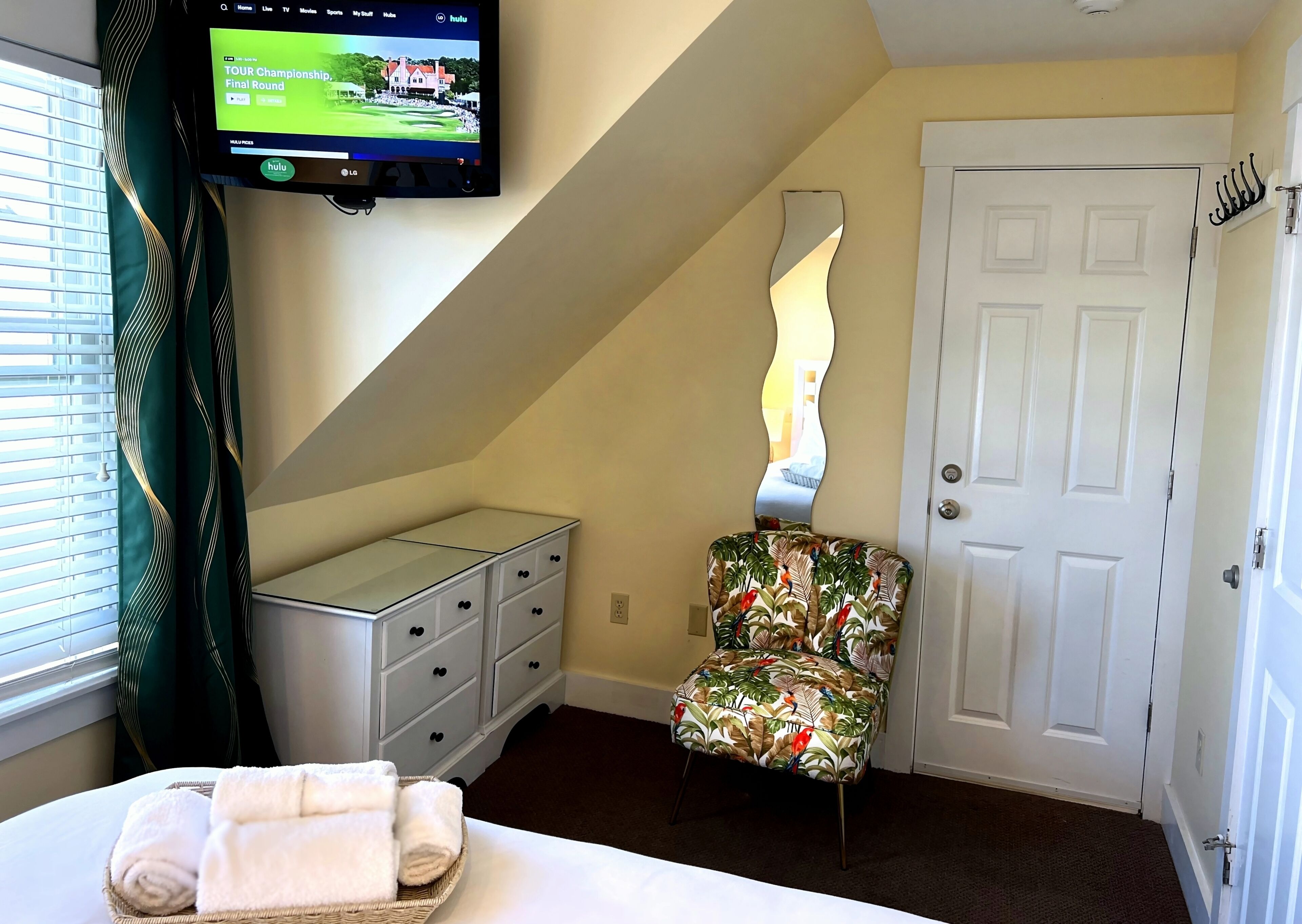 Une chambre d’hôtel avec un lit, un téléviseur fixé au mur, une porte blanche, une chaise à motifs tropicaux et une fenêtre avec des stores.