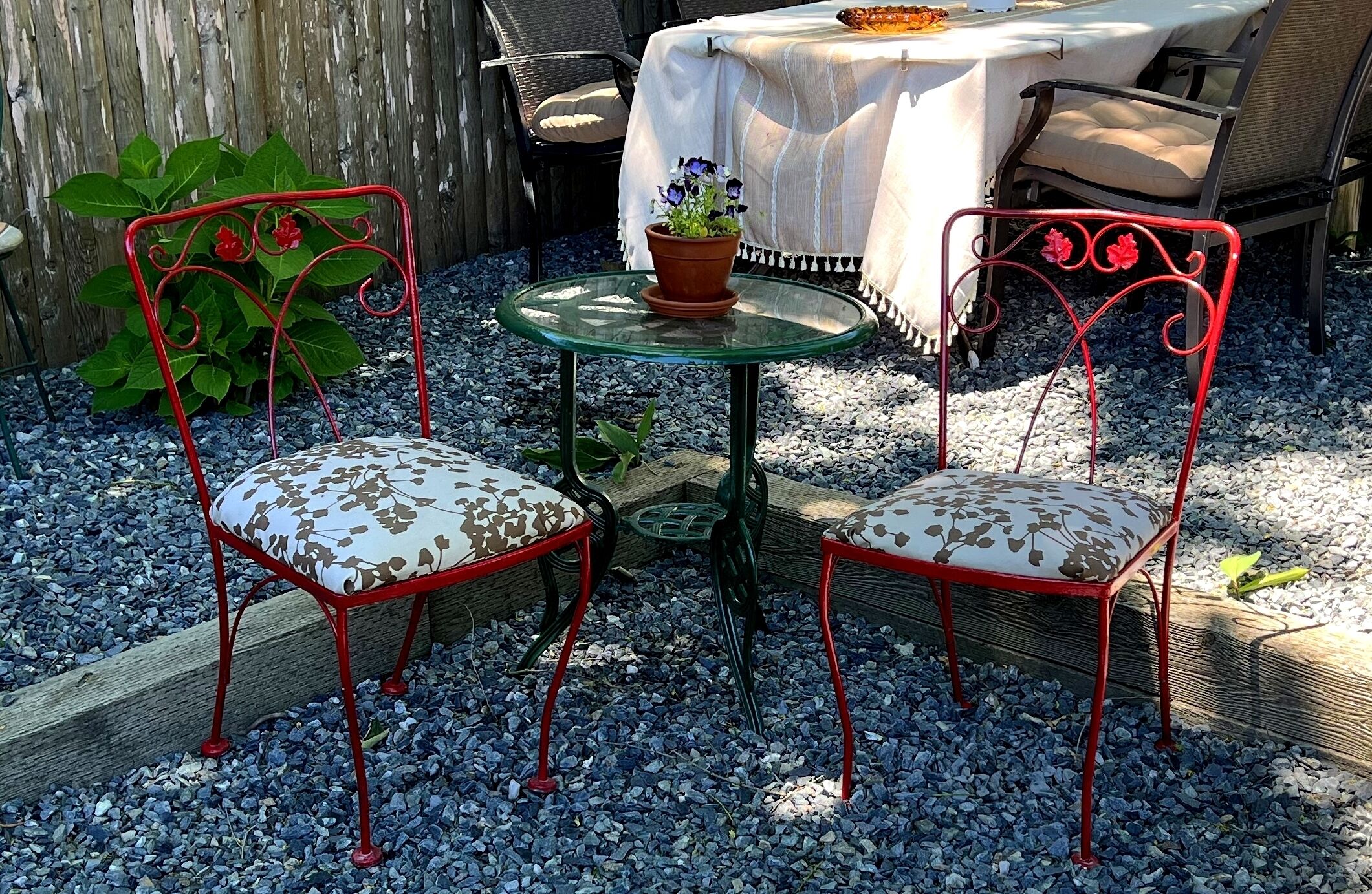 A patio area with two red metal chairs with patterned cushions, a glass table with a potted plant, and a covered table with chairs.
