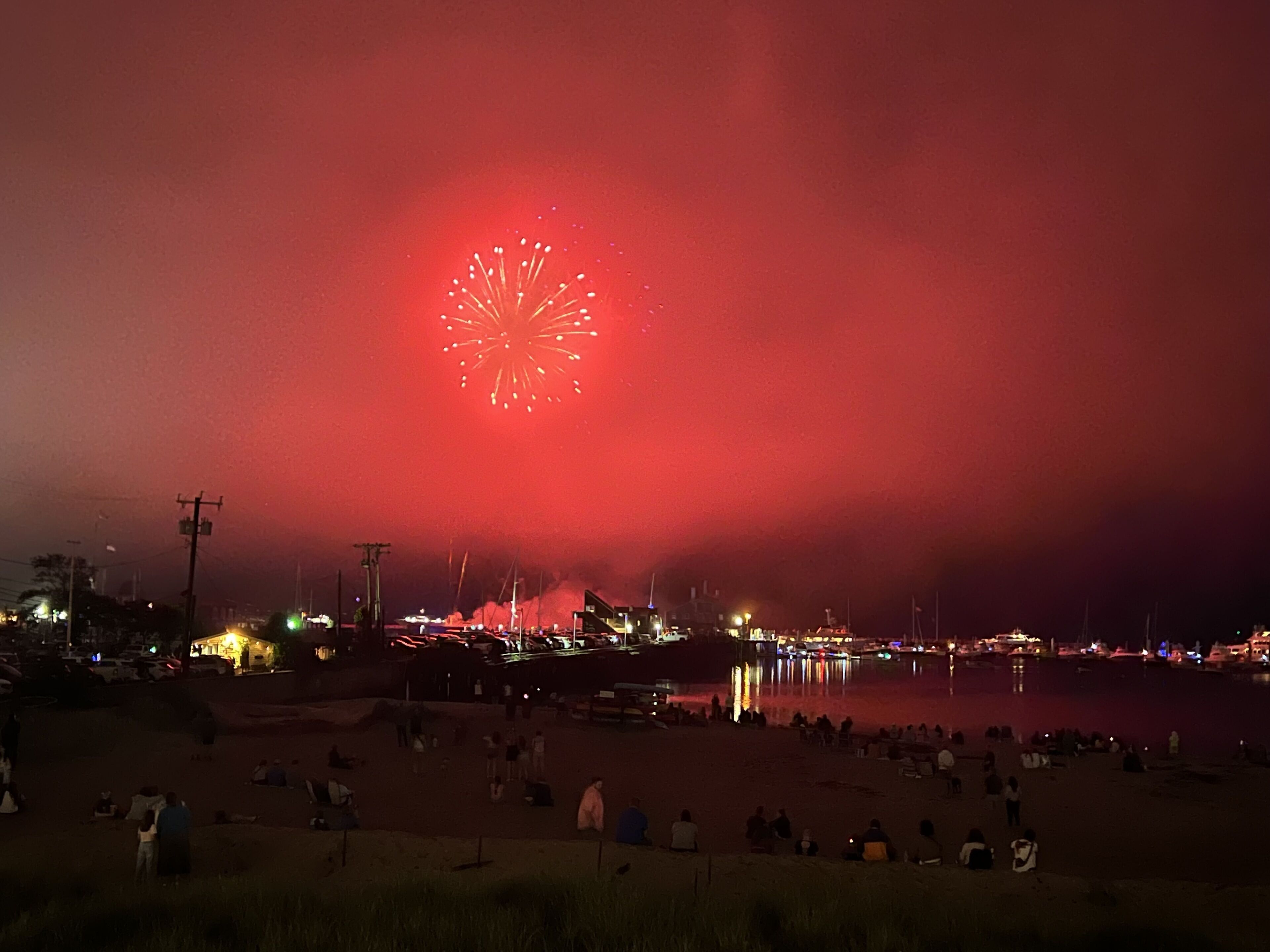 Una playa con gente sentada y de pie, y un gran espectáculo de fuegos artificiales en el cielo.