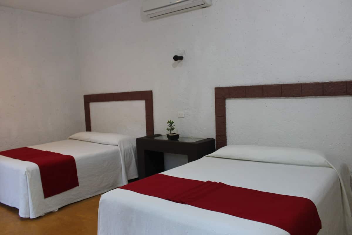 Habitación de hotel con dos camas individuales, cada una con una colcha roja, una mesita con una planta y un aire acondicionado instalado en la pared.