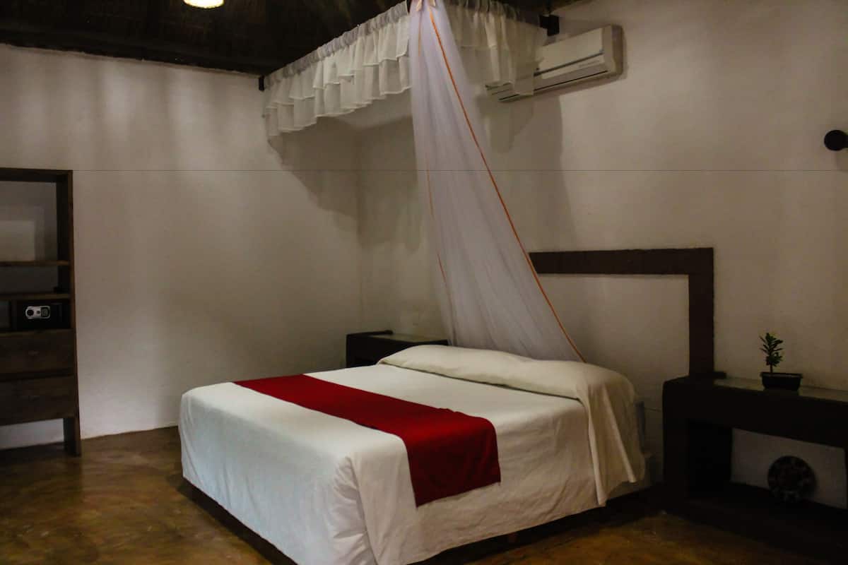 Un dormitorio con cama, una mesita de noche, una manta roja y un dosel blanco, además de un equipo de aire acondicionado en la pared.