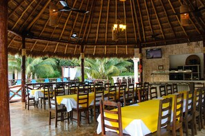 Ein Restaurant mit Strohdach, Holztischen und -stühlen mit Blick auf Palmen und einen Pool.