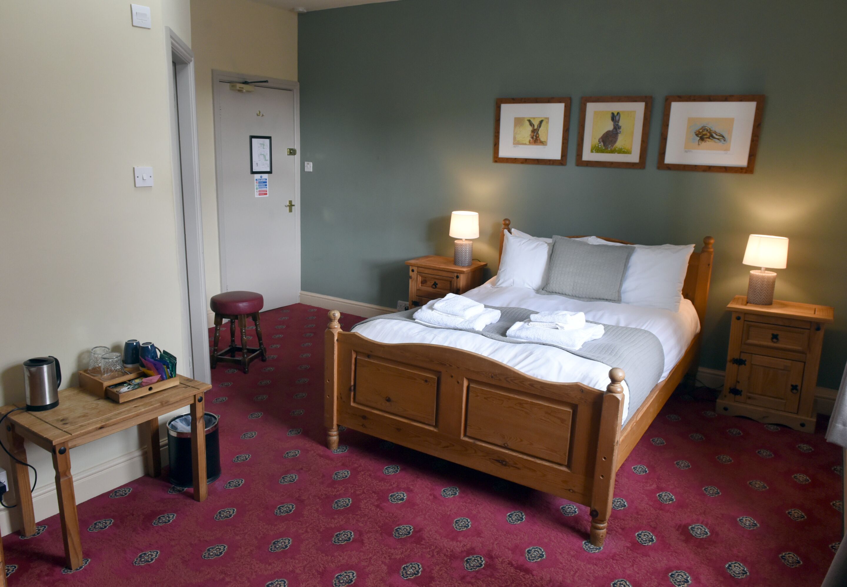 A hotel room with a wooden four-poster bed, two bedside tables with lamps, a red patterned carpet, and three framed pictures on the wall.