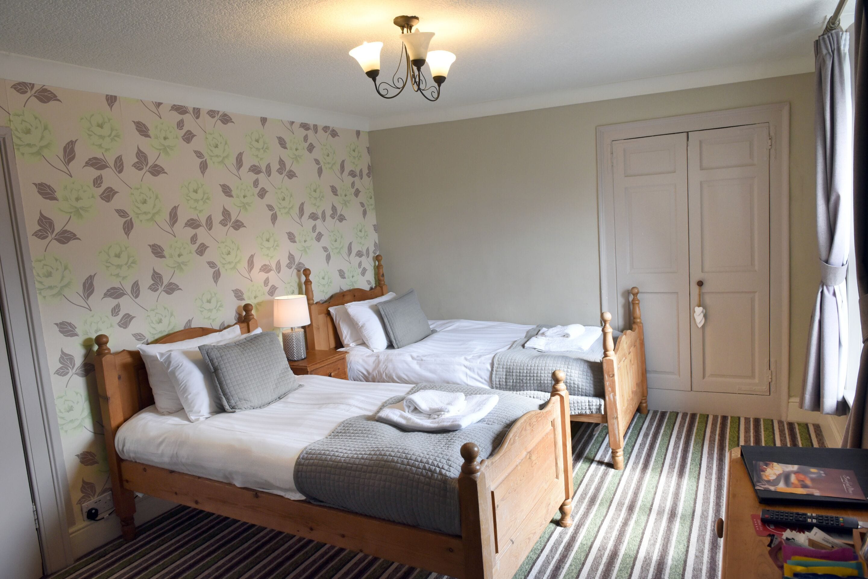 A hotel room with two wooden beds, a striped carpet, a floral wallpaper, and a white door.
