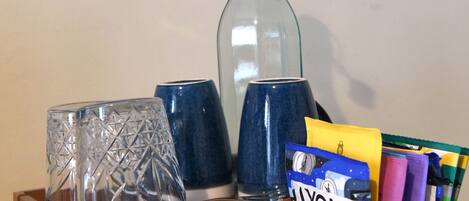 Una bandeja de madera con una botella de vidrio, dos tazas azules, un vaso con diseño de diamantes y un paquete de cigarrillos Lyons.
