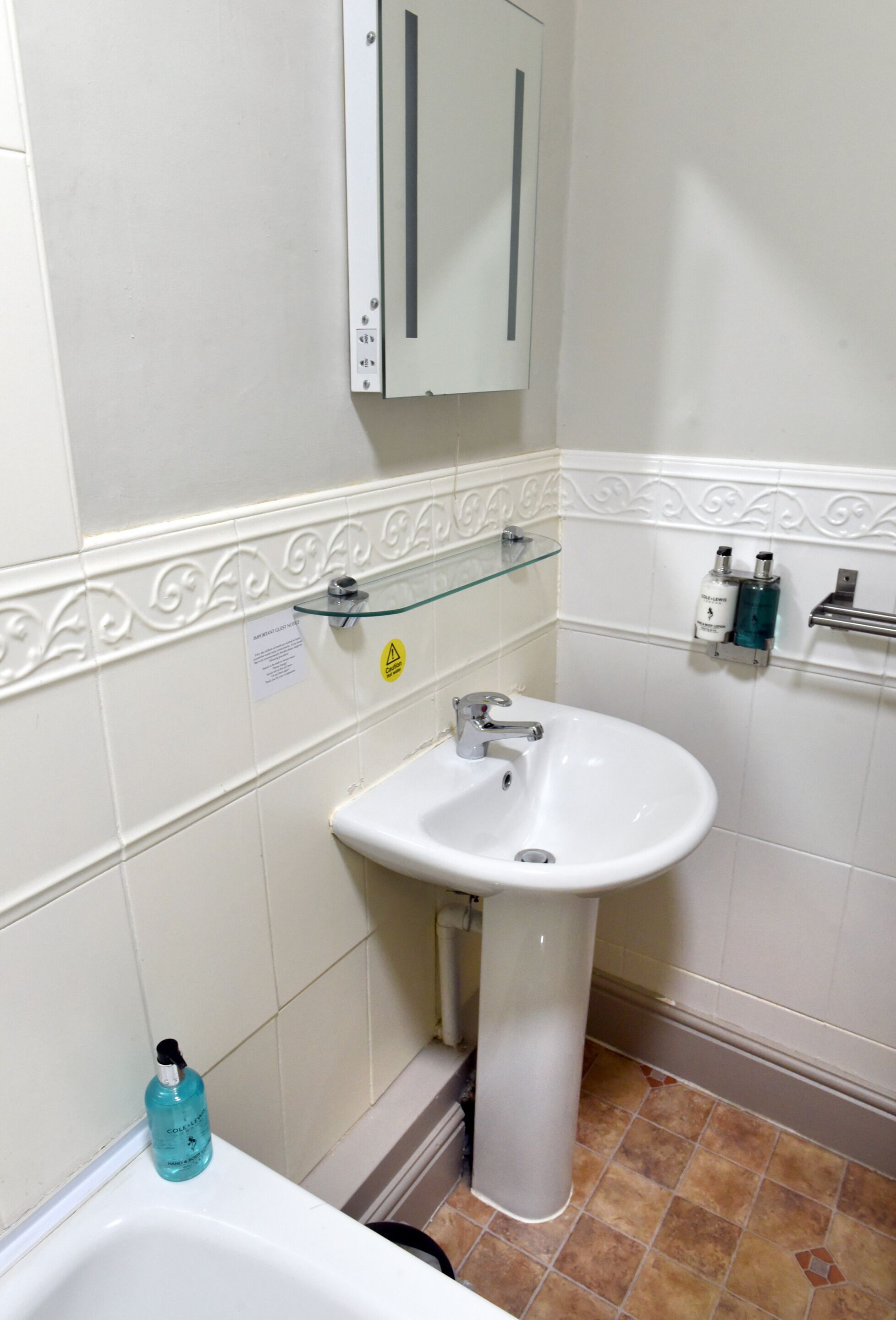 Een badkamer met een witte wasbak, een glazen plank, een kast en een handzeepdispenser.