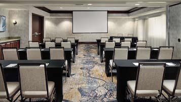 Un salón de conferencias con piso alfombrado, una pantalla de proyector y varias mesas con sillas.