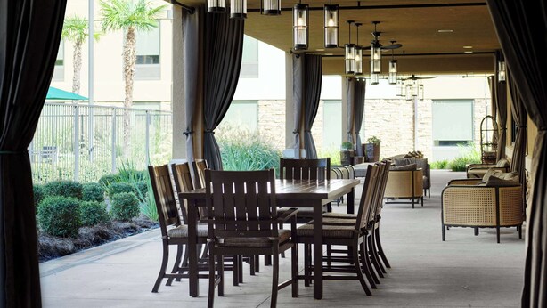 An outdoor dining area with a long table and multiple chairs, surrounded by curtains and hanging lights. - Hilton Garden Inn Bossier City, LA (Bossier City)
