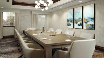 Een congrescentrum met een lange tafel, witte stoelen en een kroonluchter. Er hangen drie schilderijen aan de muur en er is een raam met gordijnen.