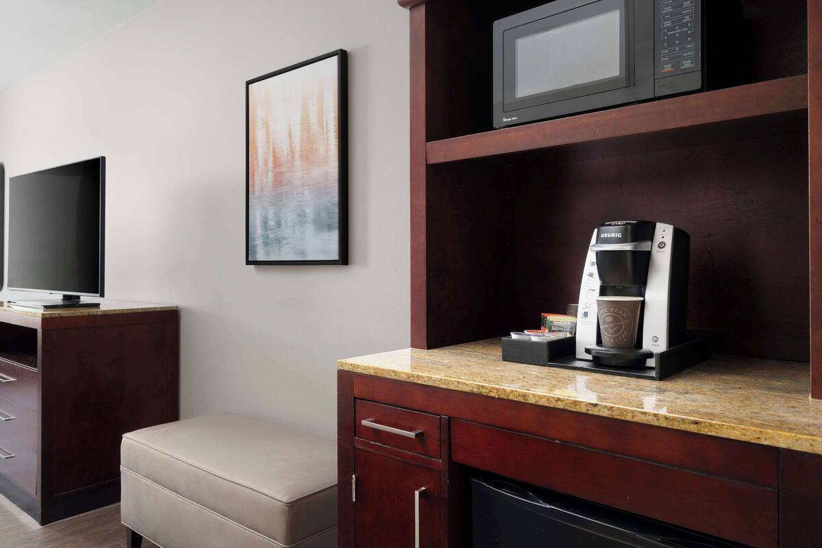 A hotel room with a flat-screen TV, a framed abstract painting, a coffee maker, and a microwave on the counter.
