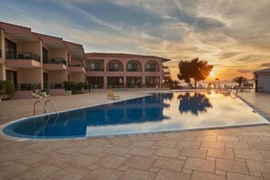 A swimming pool with a clear view of the sunset, surrounded by a building with balconies and a tiled patio area. - Toroni Blue Sea Hotel & Spa (Sithonia)
