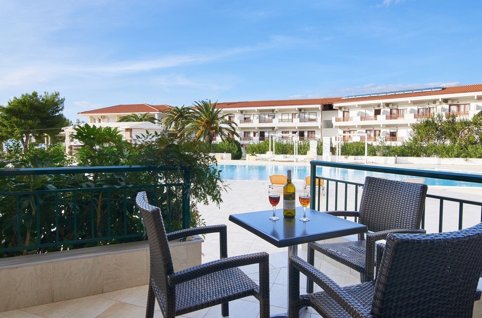 A balcony with wicker chairs and a table, overlooking a swimming pool and a building with red-tiled roofs.