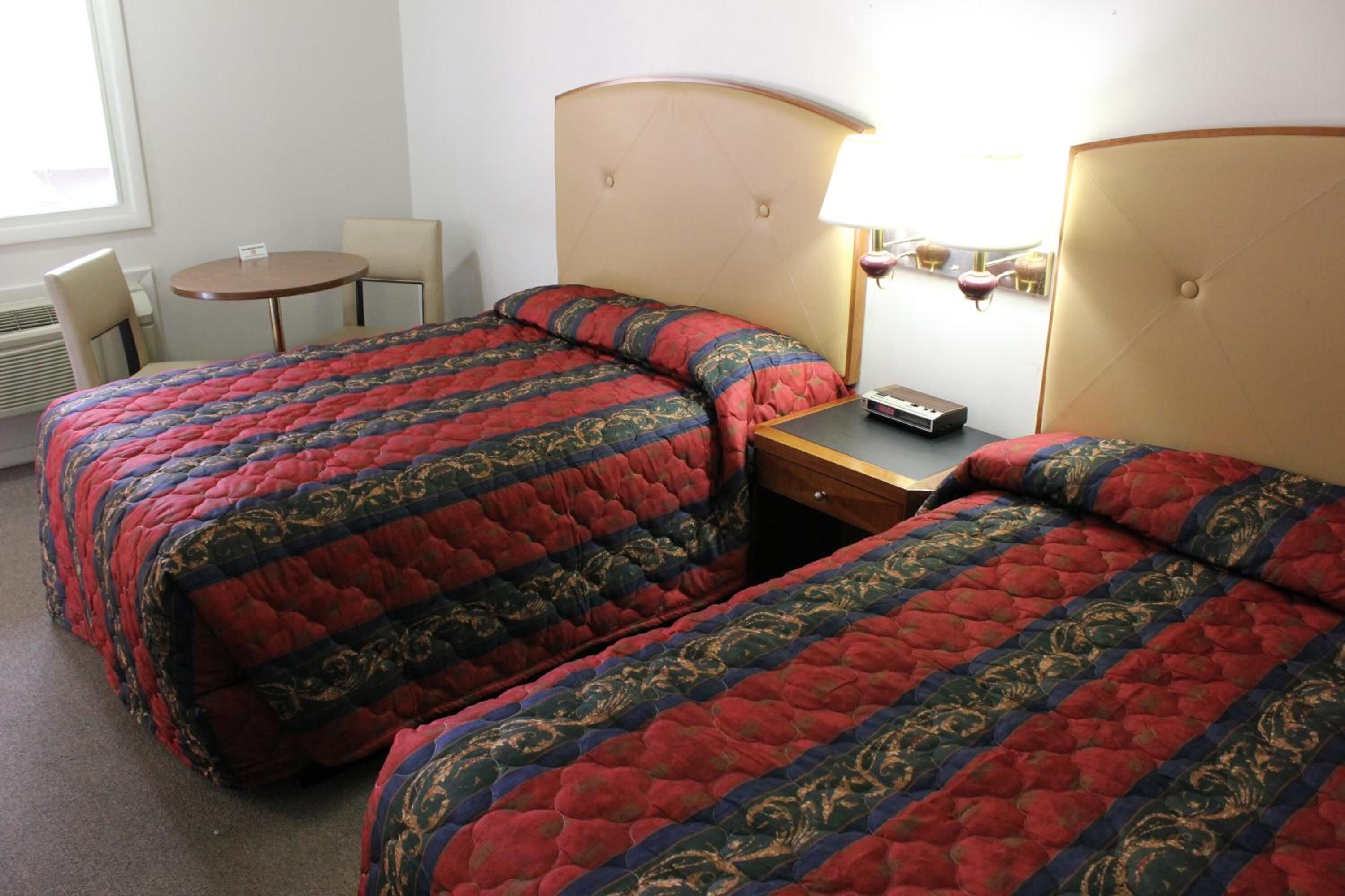 Quarto de hotel com duas camas, uma mesinha, uma cadeira e um abajur.