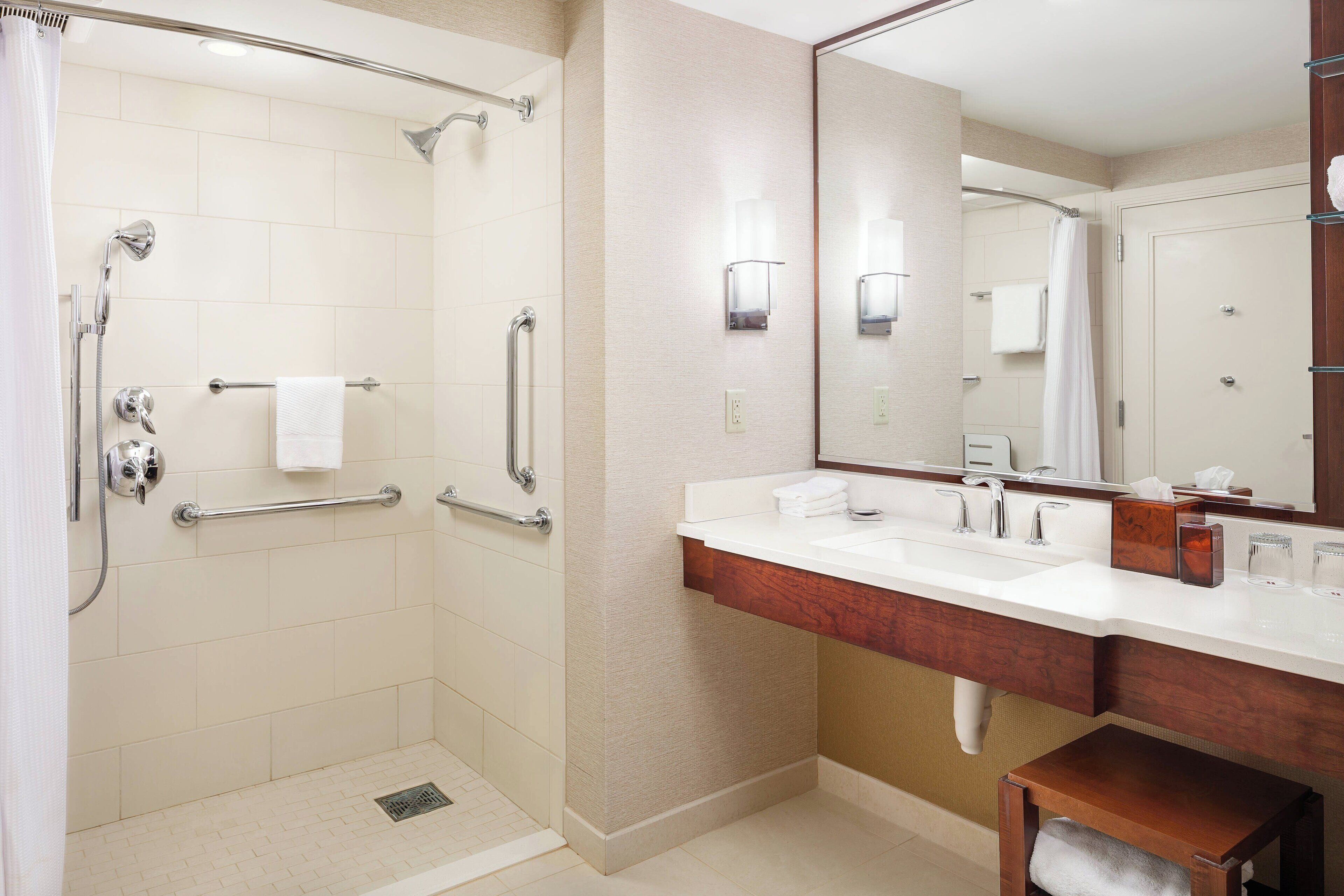 Ein modernes Badezimmer mit bodengleicher Dusche, Haltegriffen, einem großen Spiegel und einem Waschtisch mit Spüle und Arbeitsplatte.