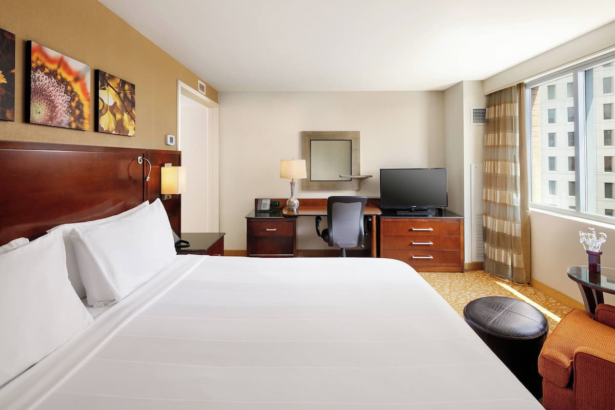 Ein Hotelzimmer mit einem großen Bett, einem Schreibtisch mit Stuhl, einem Fernseher und einem Fenster mit Vorhängen.
