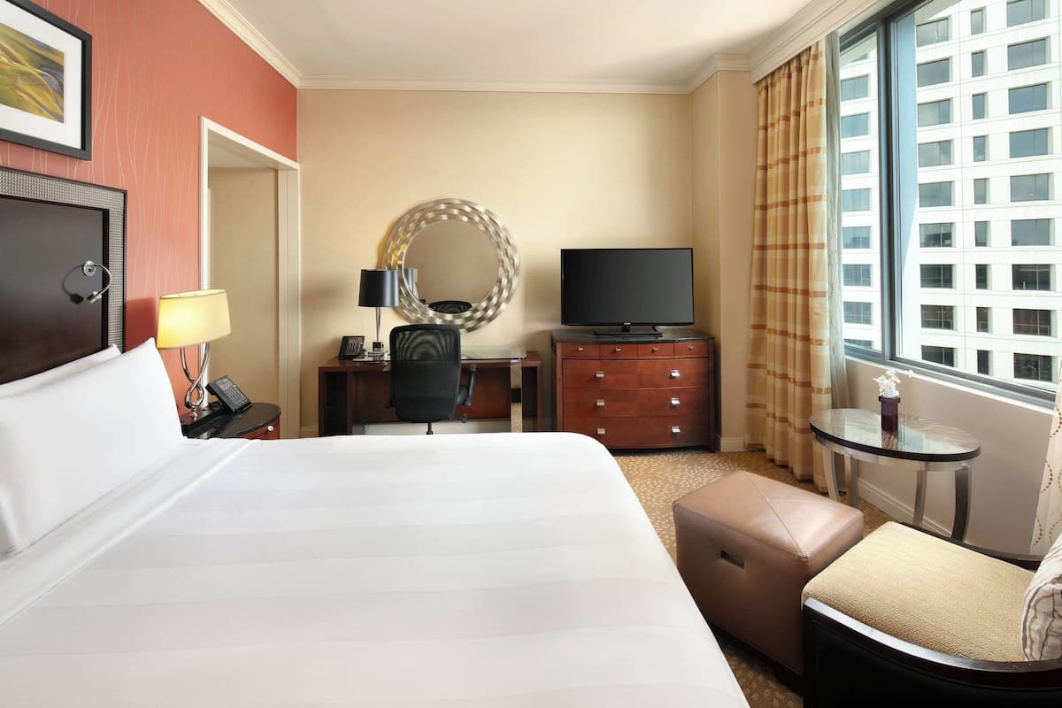 Ein Hotelzimmer mit einem großen Bett, einem Schreibtisch mit Computer, einem Fernseher, einem Spiegel, einem kleinen Tisch und einem Fenster mit Vorhängen.