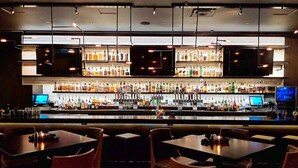 Zona de bar con varias mesas y sillas, una barra bien surtida con diferentes botellas y altavoces colgantes.