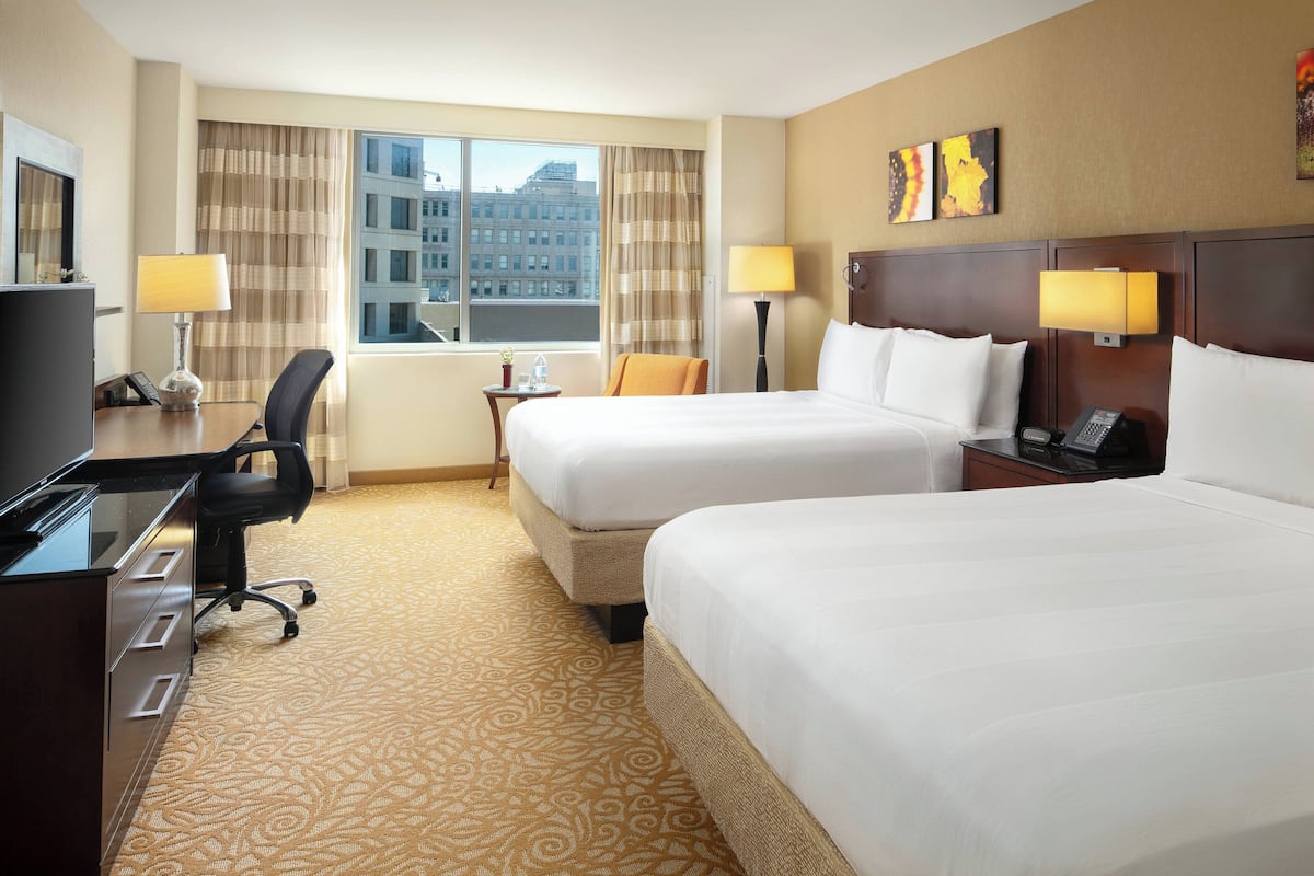 Ein Hotelzimmer mit zwei Betten, einem Schreibtisch mit Computer, einem Sessel, einem kleinen Tisch mit Lampe und Blick auf die Stadt durch große Fenster.