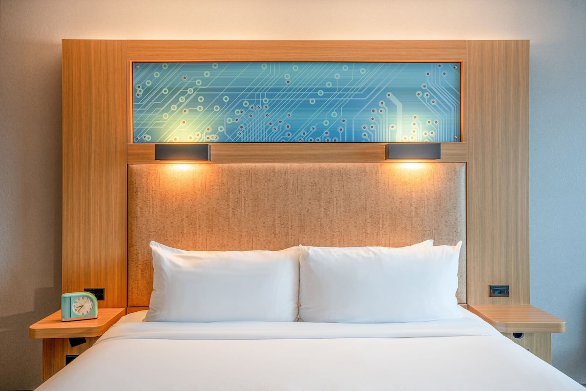 A modern hotel room with a wooden headboard, a bed with white linens, and a nightstand with a clock.