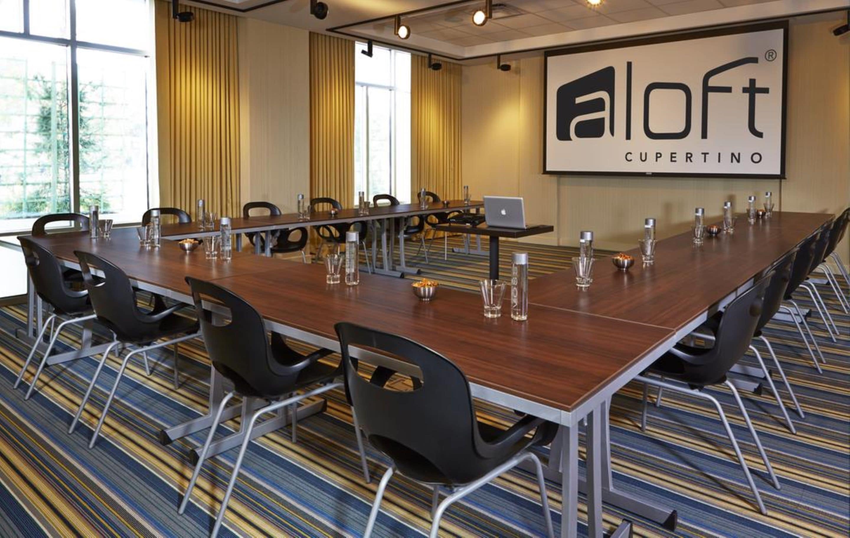 Ett konferensrum med ett långt bord, stolar och en bärbar dator. Rummet har ett stort fönster med gardiner, ett mattbeklätt golv och en skylt på väggen.