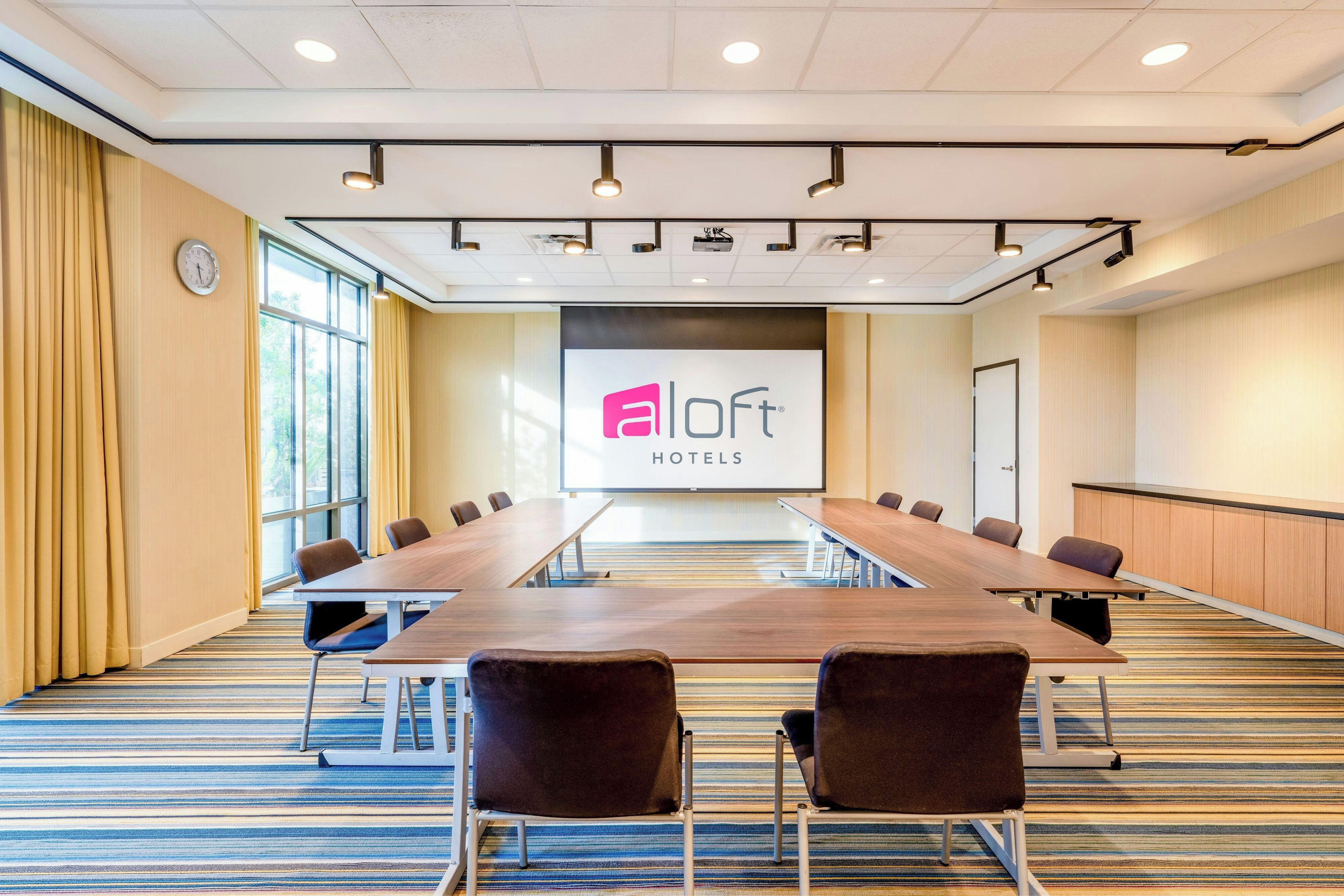 Un salón de conferencias con una larga mesa, sillas y una pantalla de presentación con el logotipo de 'aLoft Hotels'.