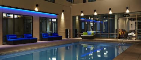Una piscina interior con zonas de estar azules y blancas, rodeada por paredes de vidrio e iluminación moderna.