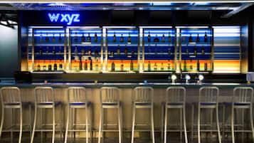 Zona de bar con un letrero de neón que dice "WXYZ" y varias estanterías llenas de botellas. Hay seis taburetes de bar frente a ellas.