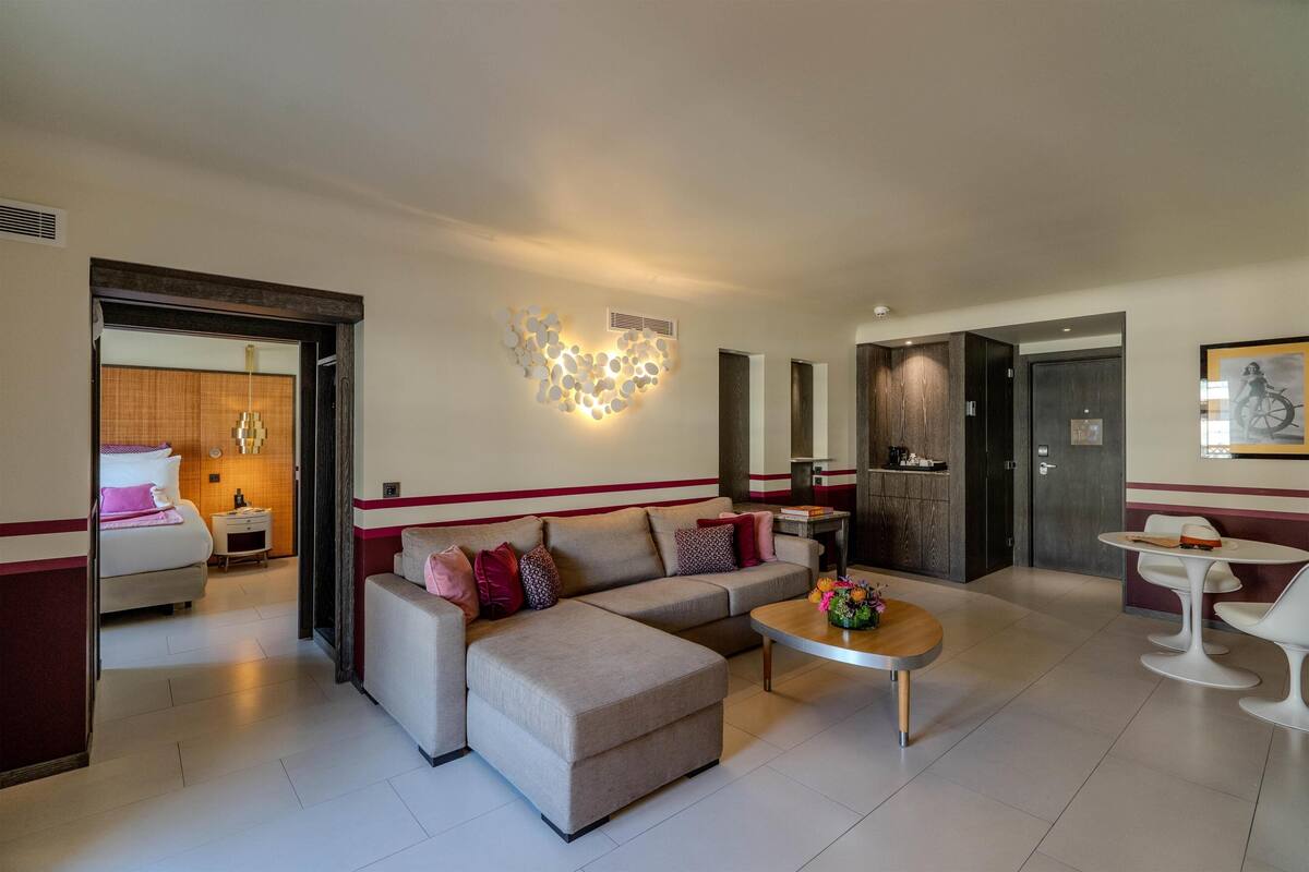 Une chambre d’hôtel moderne équipée d’un canapé, d’un pouf, d’une table basse et d’un coin repas avec des chaises.