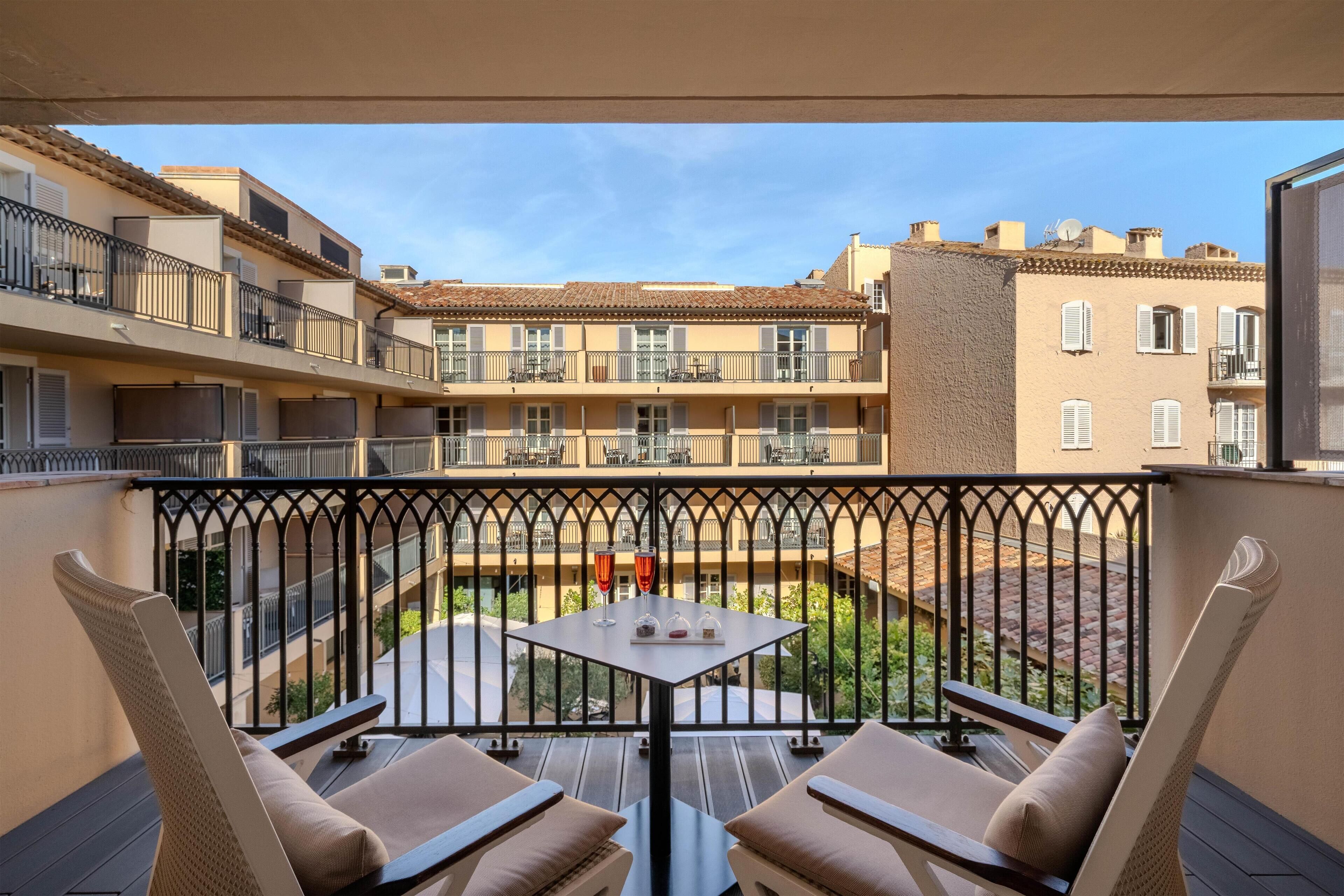 A balcony with two chairs and a table, overlooking a courtyard with buildings in the background.
