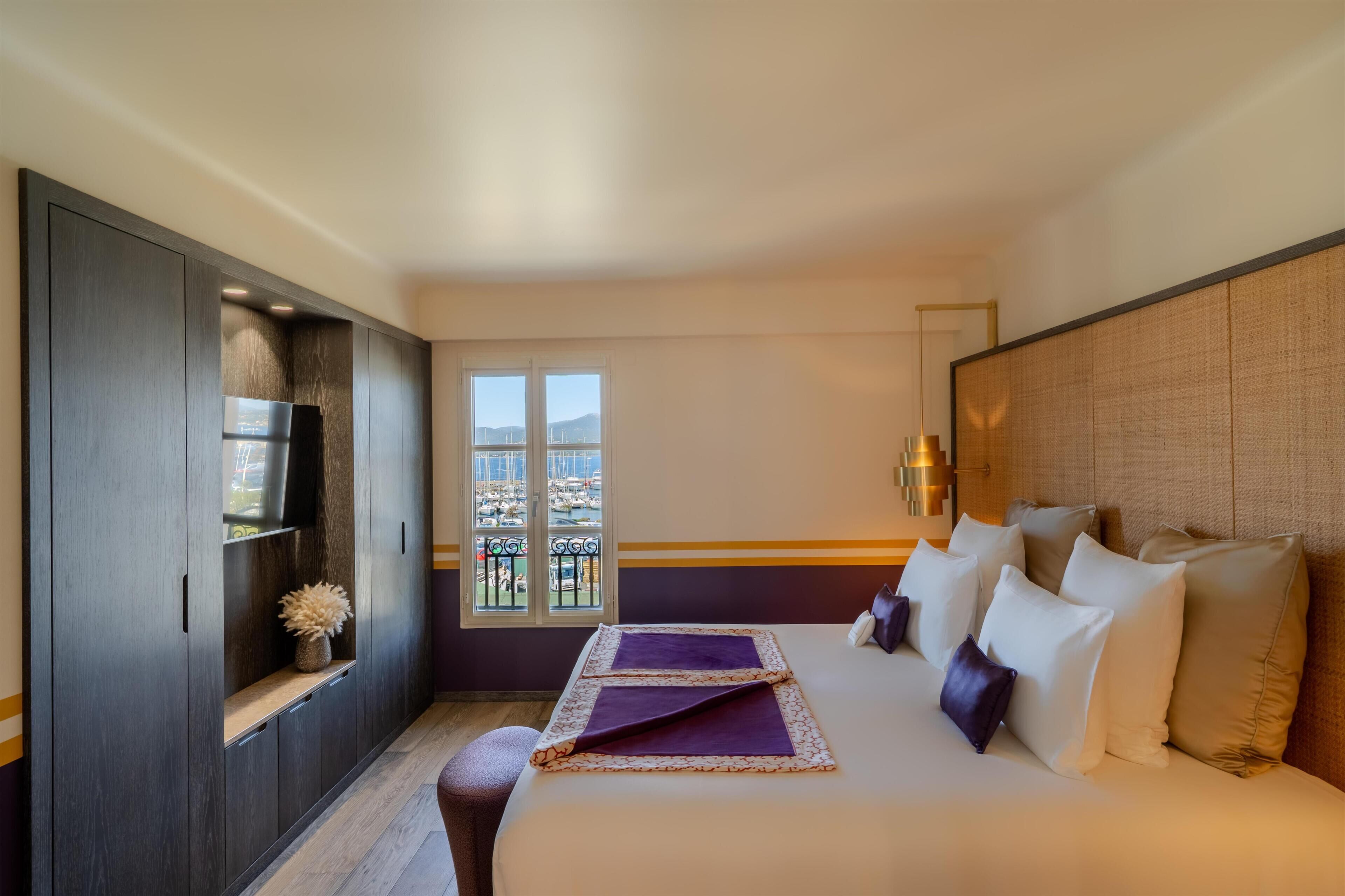 A hotel room with a bed, a wardrobe, a window with a view of boats, and a balcony.