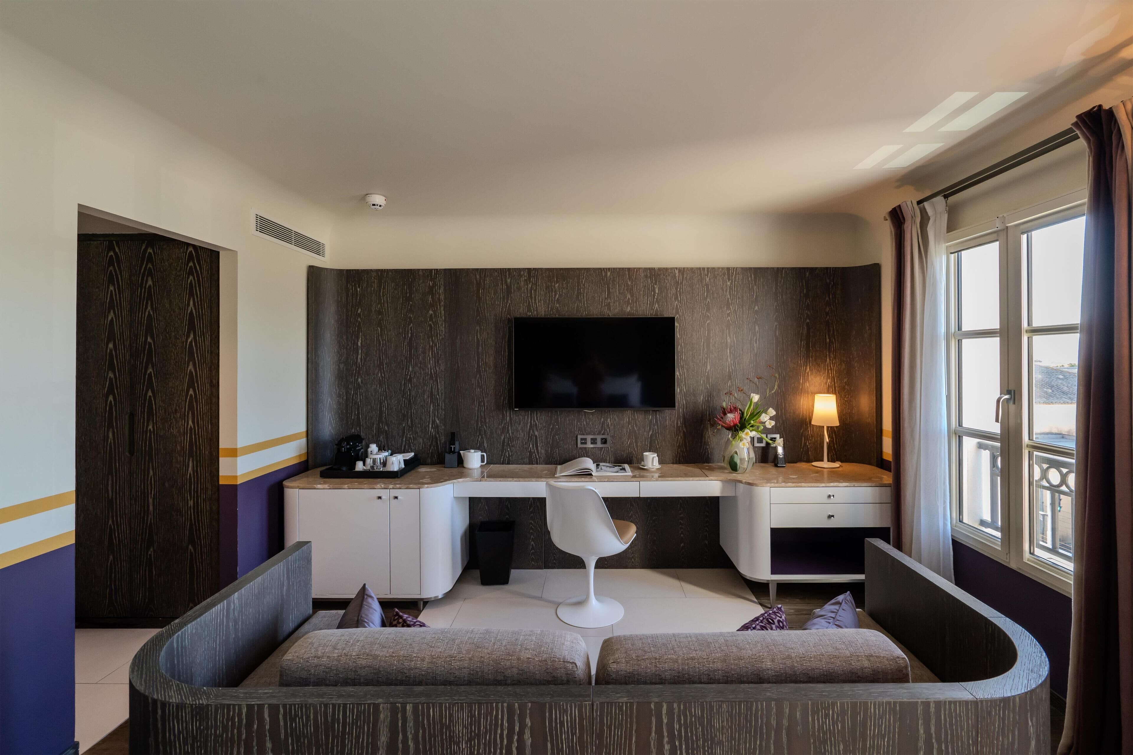 Habitación de hotel moderna con sofá curvo, escritorio con silla, televisor de pantalla plana y ventana con cortinas.