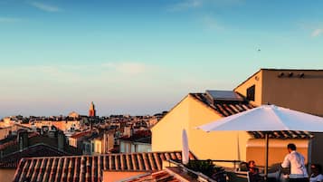 A rooftop terrace with a white umbrella, tiled roofs, and a view of a cityscape with buildings and a tower.