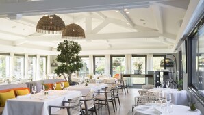 A spacious dining area with multiple tables and chairs, large windows, and hanging wicker lamps.