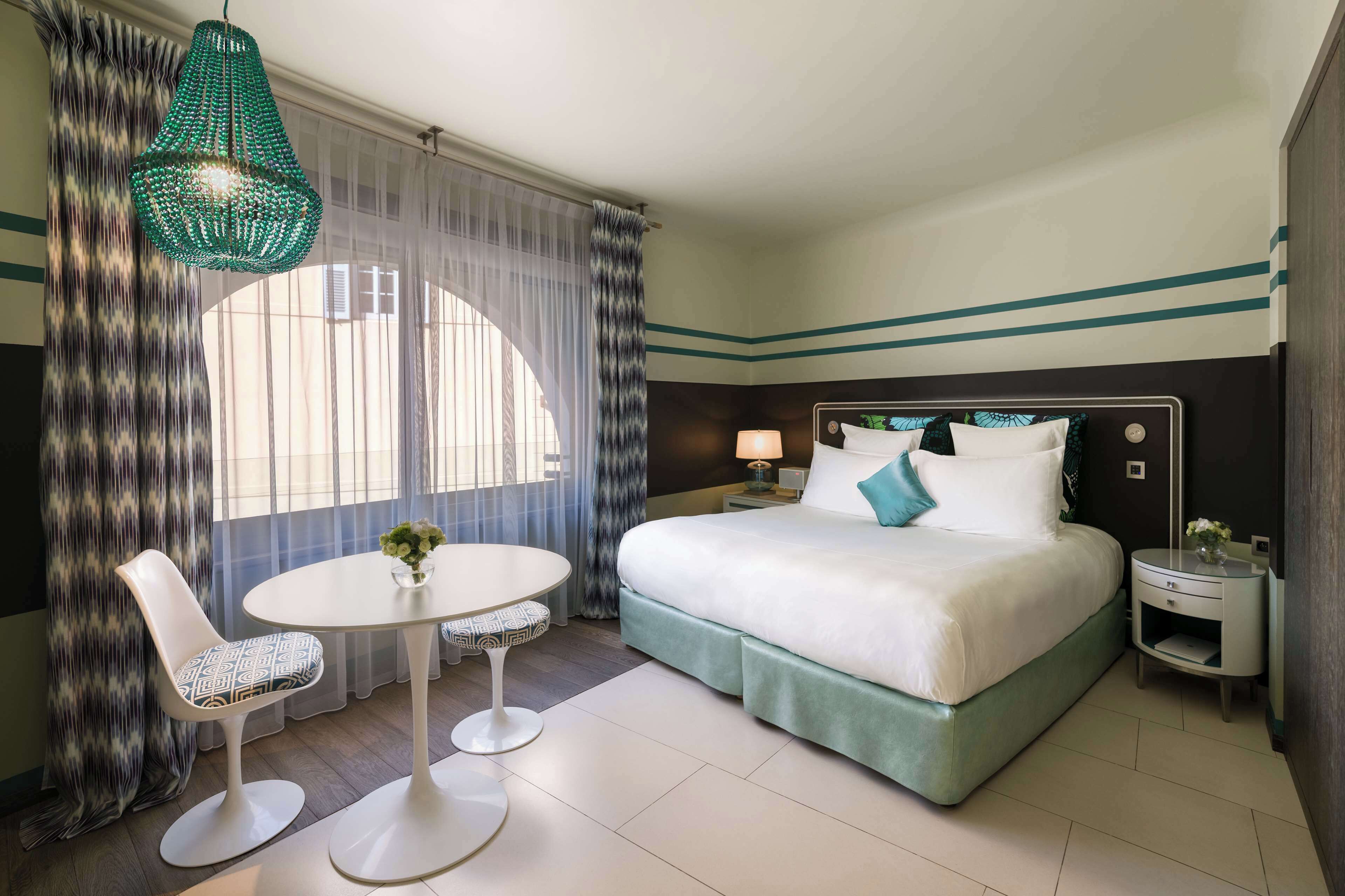 Habitación de hotel con cama, una mesa redonda con dos sillas, lámpara colgante y ventana con cortinas traslúcidas.