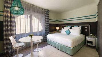 Habitación de hotel con cama, una mesa redonda con dos sillas, lámpara colgante y ventana con cortinas traslúcidas.