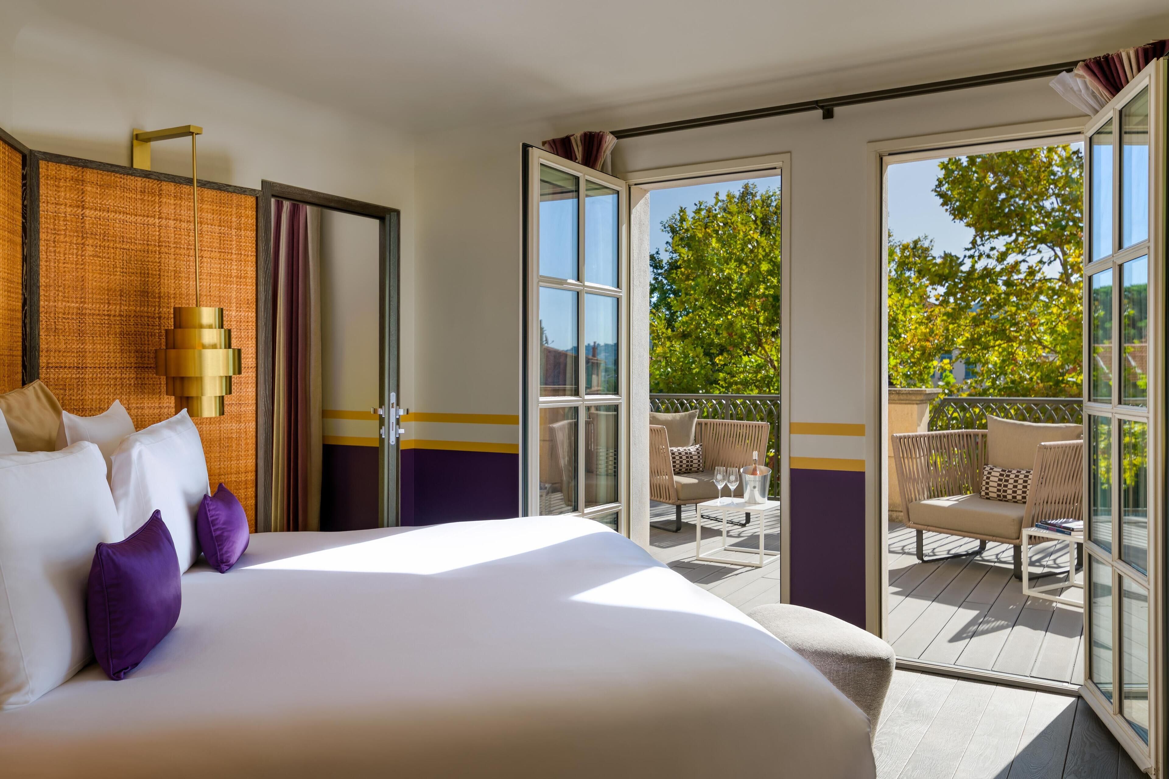 A hotel room with a large bed, a balcony with outdoor seating, and a view of greenery.