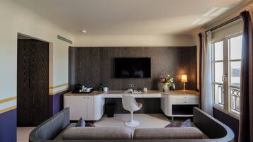 A modern hotel room with a curved sofa, a desk with a chair, a flat-screen TV, and a window with curtains.