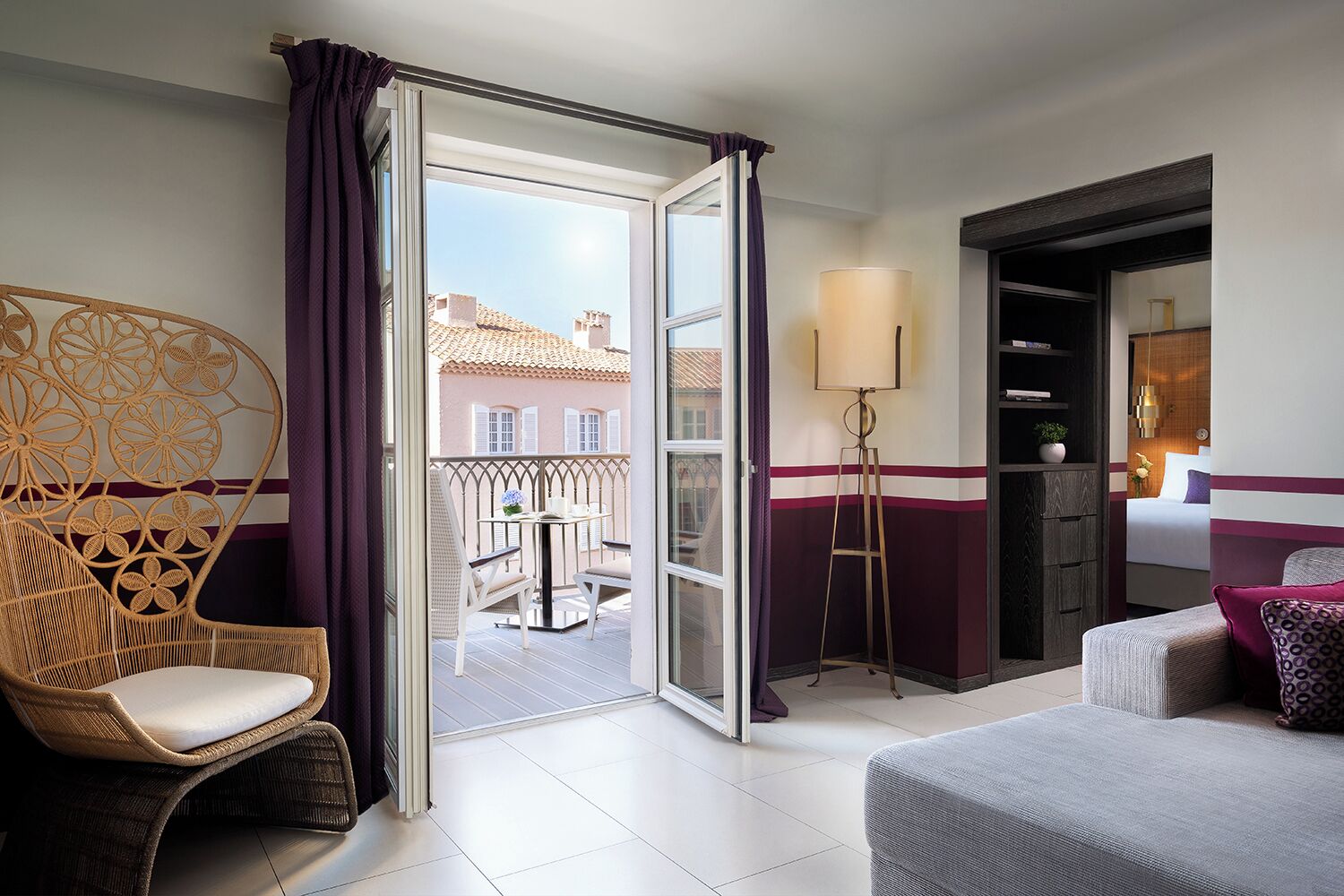 A hotel room with a balcony, a wicker chair, a floor lamp, and a bed with a patterned pillow.