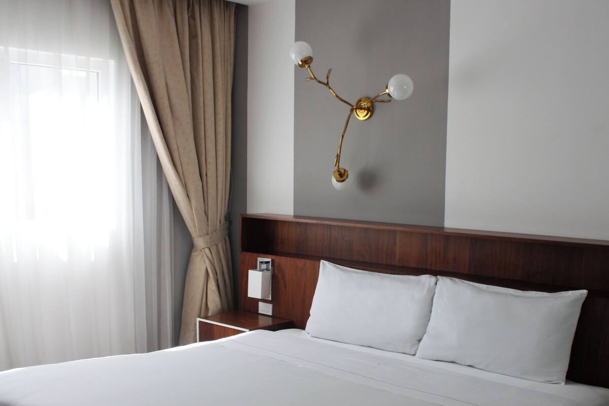 A hotel room with a large bed, a wooden headboard, a nightstand, a wall-mounted lamp, and a window with curtains.