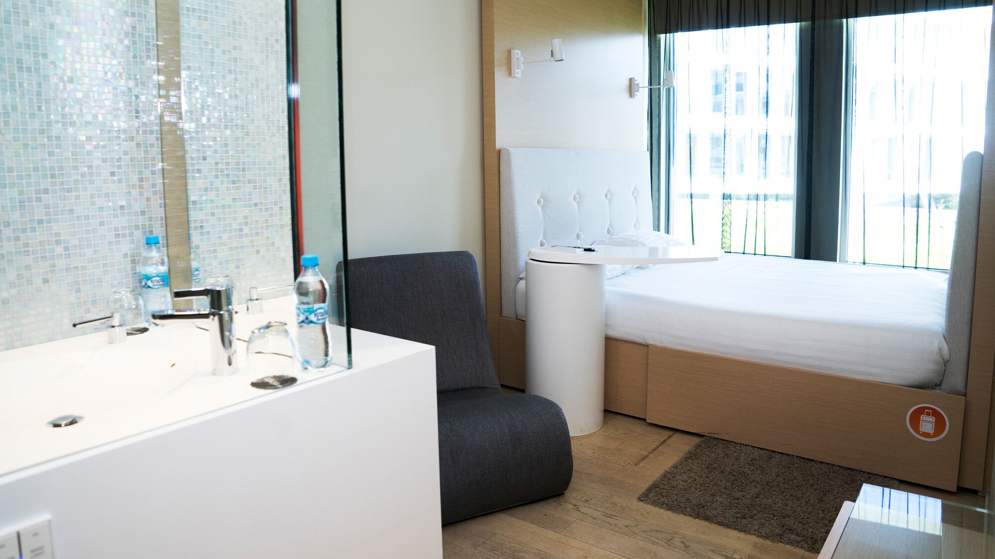 Una habitación de hotel moderna con un espejo grande, un sillón gris, una cama con cabecera de madera, una mesita de noche pequeña y una ventana con cortinas.