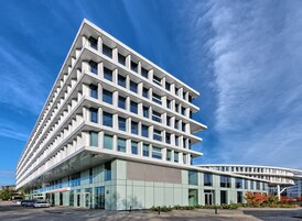 Egy modern épület, üveg homlokzattal és több szinttel, melynek feltűnő bejárata van, és egy tábla is található rajta.