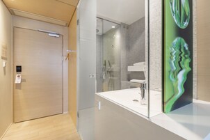 A modern bathroom with a glass shower enclosure, a white sink, and a mirror with a green abstract design.