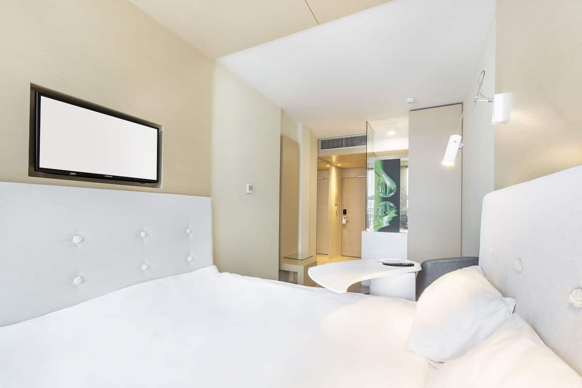 A modern hotel room with a large bed, a small desk, a mirror, and a bathroom visible through an open door.