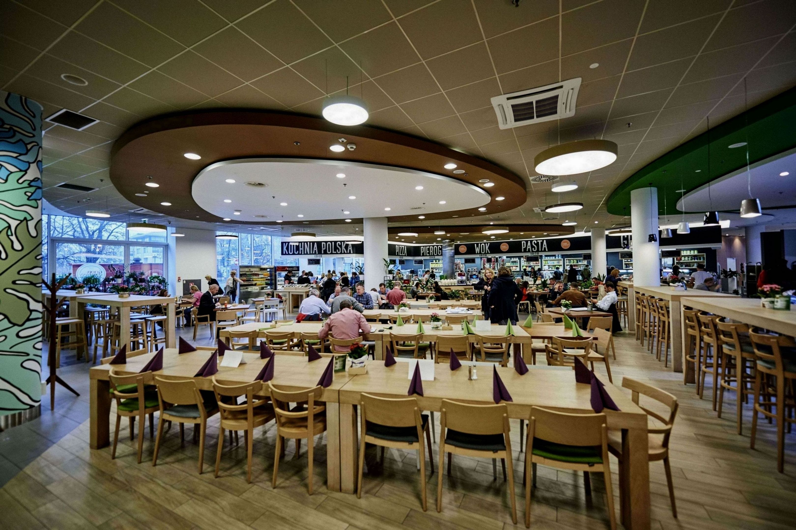 A spacious cafeteria with multiple dining tables and chairs, featuring a variety of food outlets in the background.