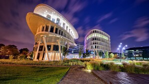 Un edificio moderno con una arquitectura curva distintiva, rodeado de un jardín cuidado y farolas.